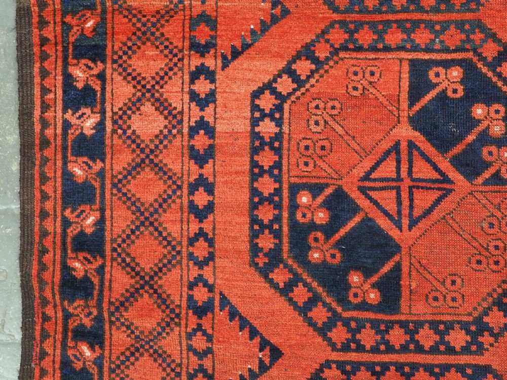 An antique handwoven Ersari Turkmen carpet featuring repeating octagonal guls in dark navy and red set upon a vibrant terracotta red background with geometric borders. View VII