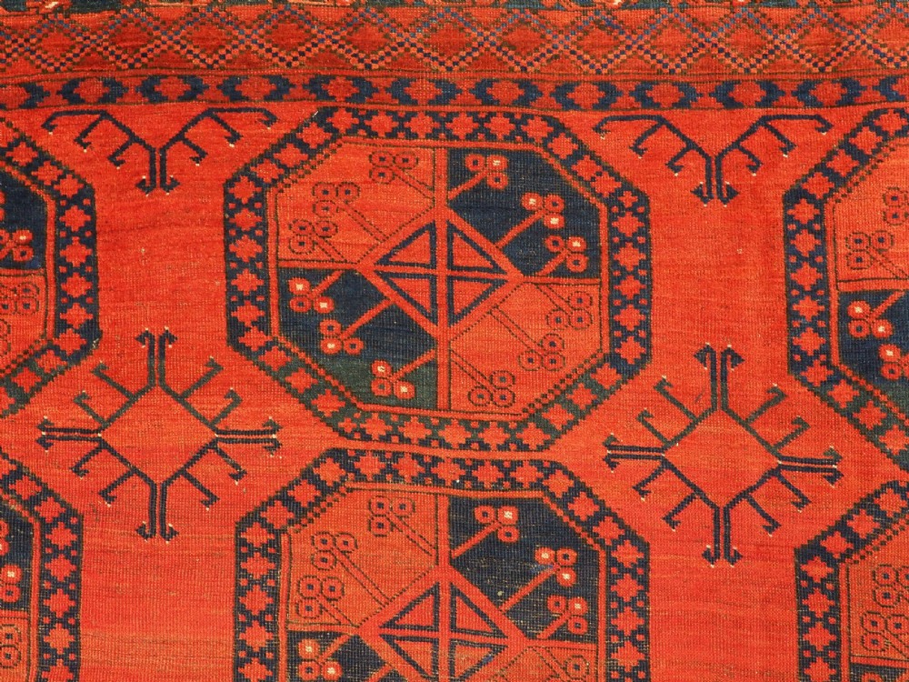 An antique handwoven Ersari Turkmen carpet featuring repeating octagonal guls in dark navy and red set upon a vibrant terracotta red background with geometric borders. View VIII