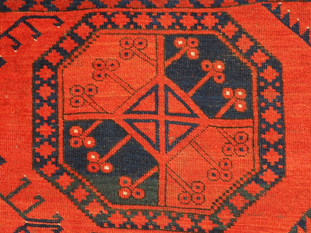 An antique handwoven Ersari Turkmen carpet featuring repeating octagonal guls in dark navy and red set upon a vibrant terracotta red background with geometric borders. View IX