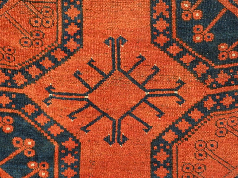 An antique handwoven Ersari Turkmen carpet featuring repeating octagonal guls in dark navy and red set upon a vibrant terracotta red background with geometric borders. View X