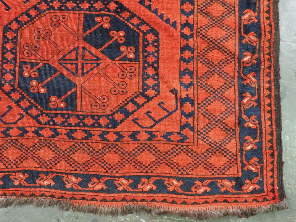 An antique handwoven Ersari Turkmen carpet featuring repeating octagonal guls in dark navy and red set upon a vibrant terracotta red background with geometric borders. View XII