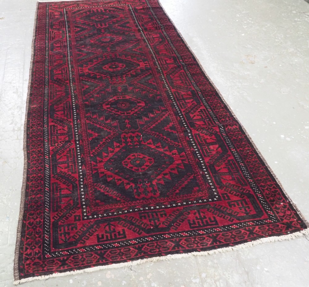 An antique Afghan Mushwani Baluch woven wool runner rug featuring deep crimson red and charcoal geometric diamond medallions along a narrow rectangular field with decorative borders. View II