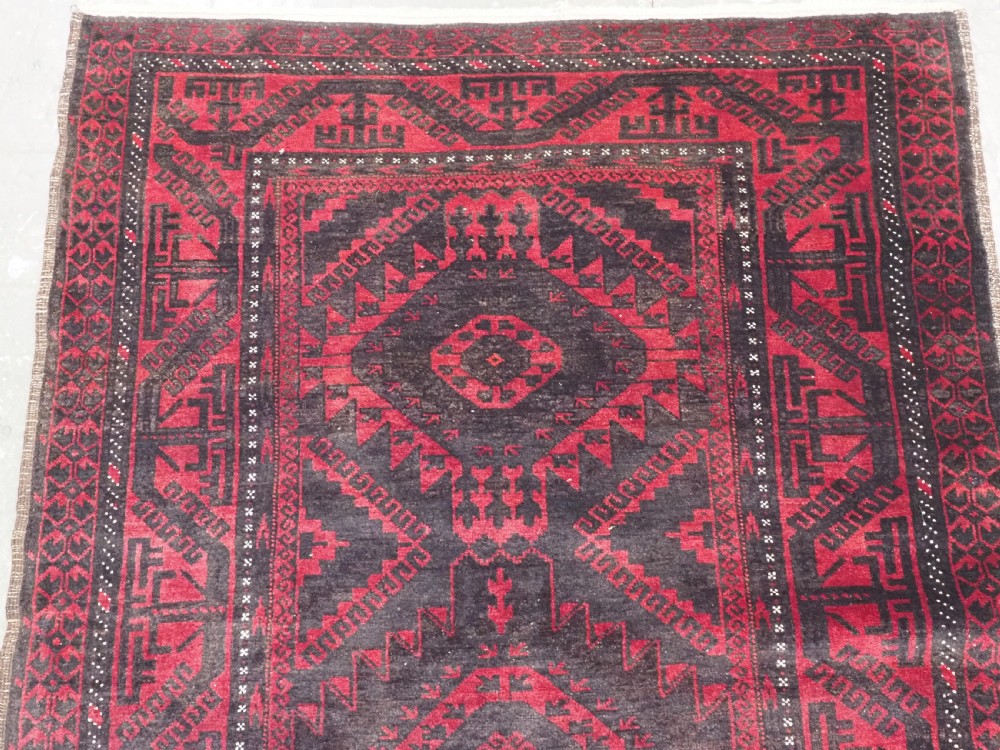 An antique Afghan Mushwani Baluch woven wool runner rug featuring deep crimson red and charcoal geometric diamond medallions along a narrow rectangular field with decorative borders. View III