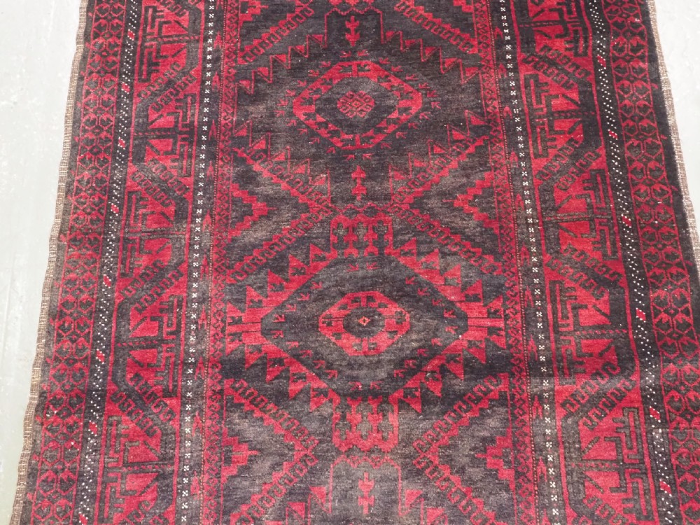 An antique Afghan Mushwani Baluch woven wool runner rug featuring deep crimson red and charcoal geometric diamond medallions along a narrow rectangular field with decorative borders. View IV