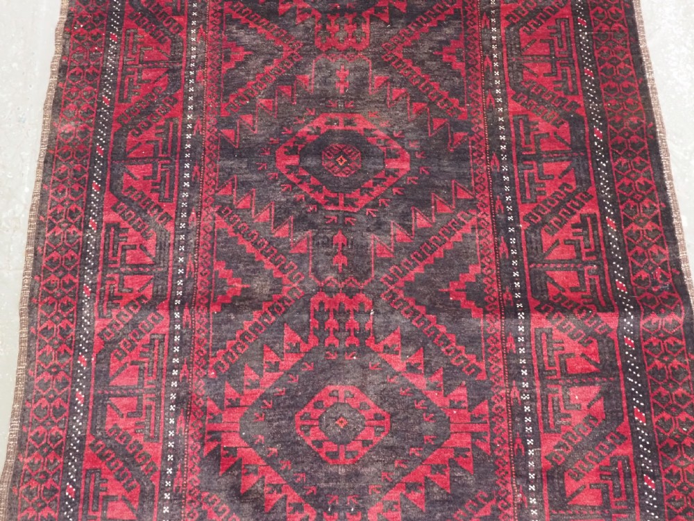 An antique Afghan Mushwani Baluch woven wool runner rug featuring deep crimson red and charcoal geometric diamond medallions along a narrow rectangular field with decorative borders. View V