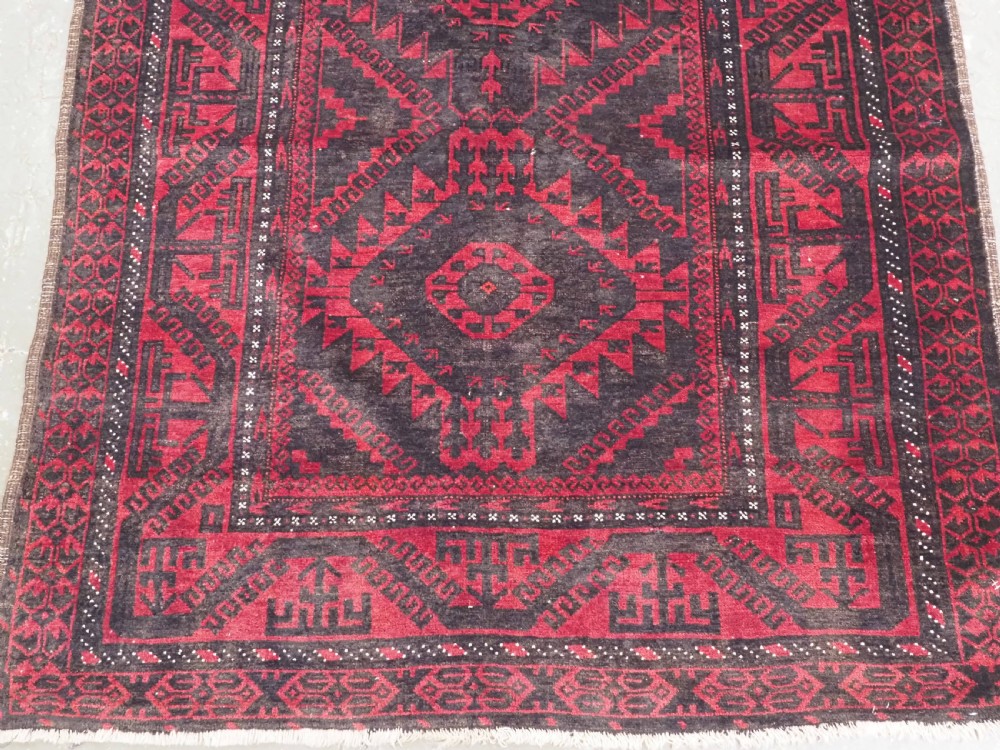 An antique Afghan Mushwani Baluch woven wool runner rug featuring deep crimson red and charcoal geometric diamond medallions along a narrow rectangular field with decorative borders. View VI