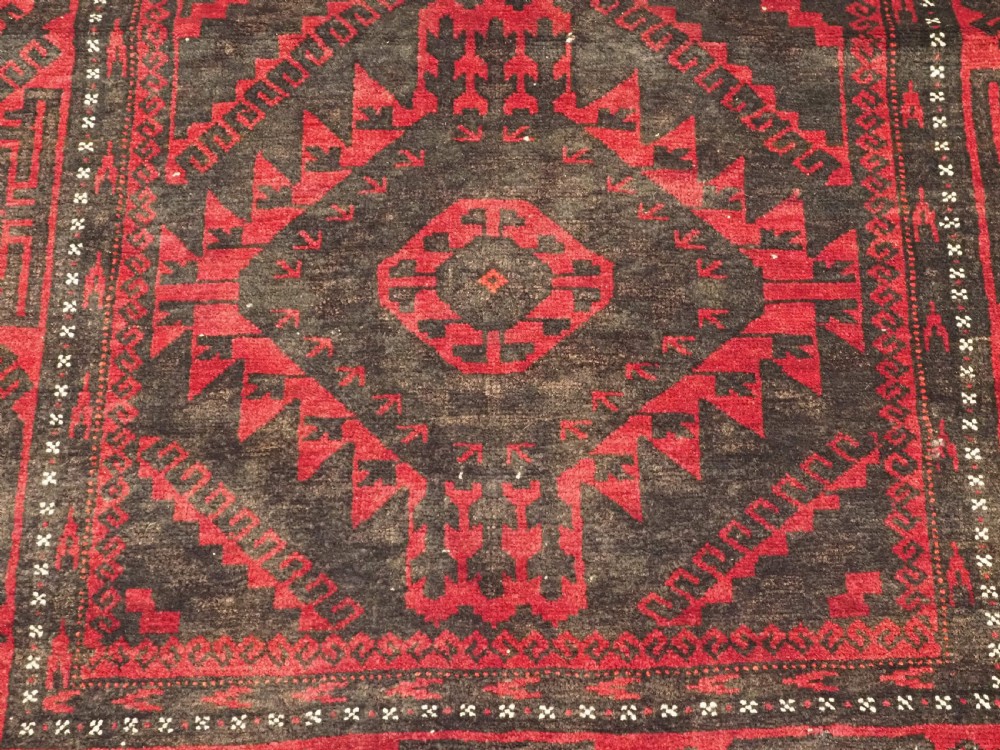 An antique Afghan Mushwani Baluch woven wool runner rug featuring deep crimson red and charcoal geometric diamond medallions along a narrow rectangular field with decorative borders. View X