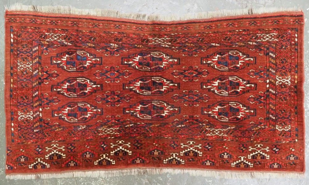 An antique rectangular Turkmen chuval wool textile featuring nine geometric gul motifs on a terracotta red ground with geometric borders.
