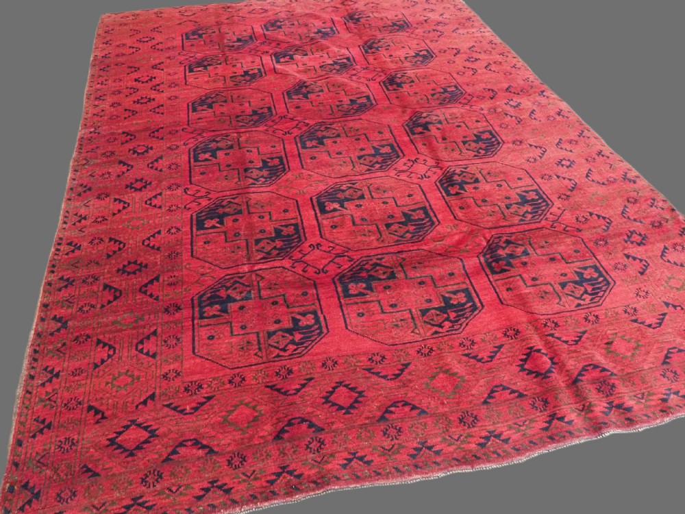 An antique rectangular wool carpet featuring a warm red field decorated with grid-arranged octagonal gul motifs within a geometric border. View II
