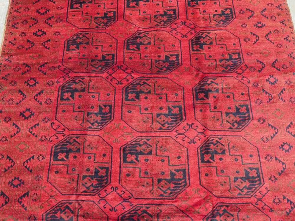 An antique rectangular wool carpet featuring a warm red field decorated with grid-arranged octagonal gul motifs within a geometric border. View IV
