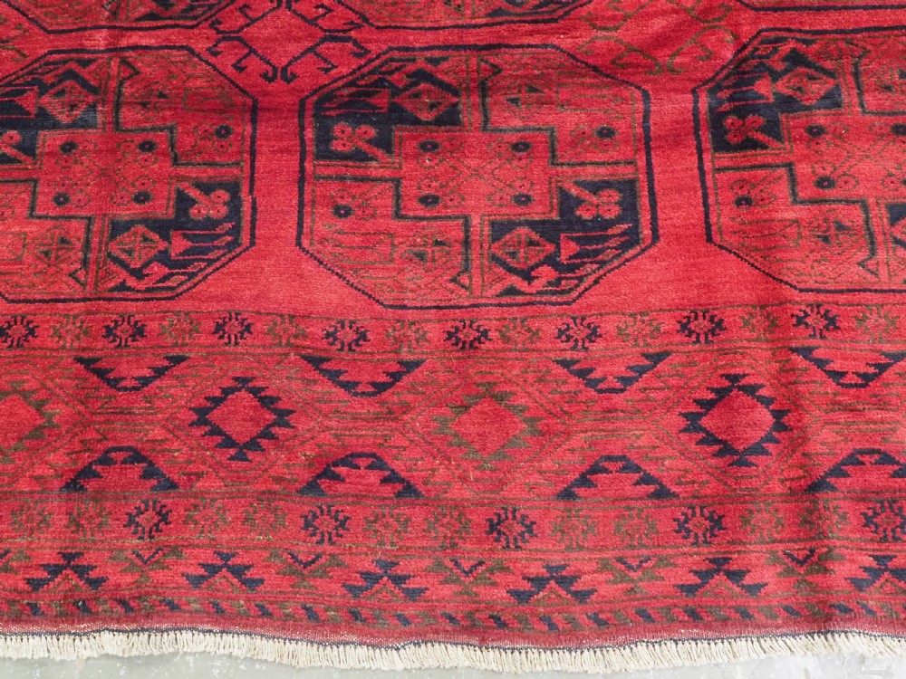 An antique rectangular wool carpet featuring a warm red field decorated with grid-arranged octagonal gul motifs within a geometric border. View X