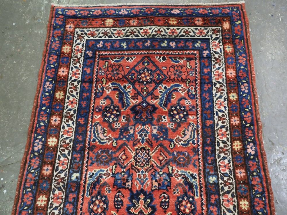 An antique long narrow Persian Kurdish wool runner rug featuring a red central field filled with geometric floral motifs framed by navy blue and cream decorative borders. View III