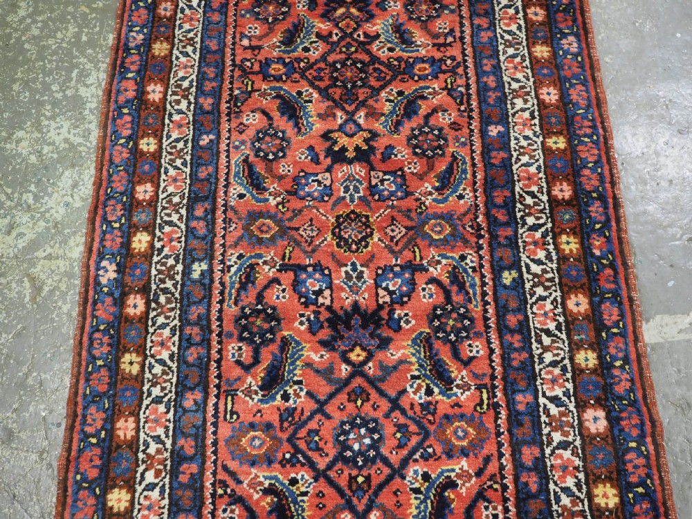 An antique long narrow Persian Kurdish wool runner rug featuring a red central field filled with geometric floral motifs framed by navy blue and cream decorative borders. View IV