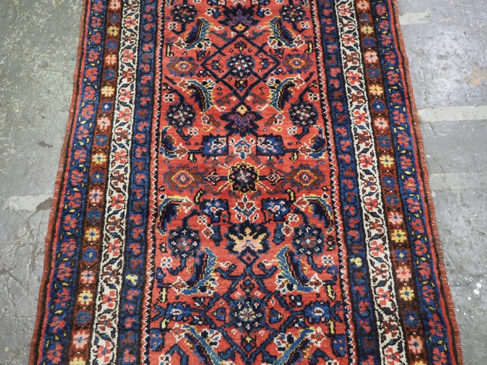 An antique long narrow Persian Kurdish wool runner rug featuring a red central field filled with geometric floral motifs framed by navy blue and cream decorative borders. View V