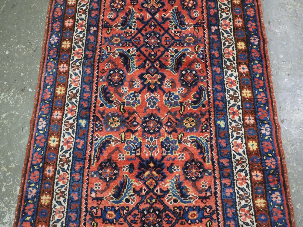 An antique long narrow Persian Kurdish wool runner rug featuring a red central field filled with geometric floral motifs framed by navy blue and cream decorative borders. View VI