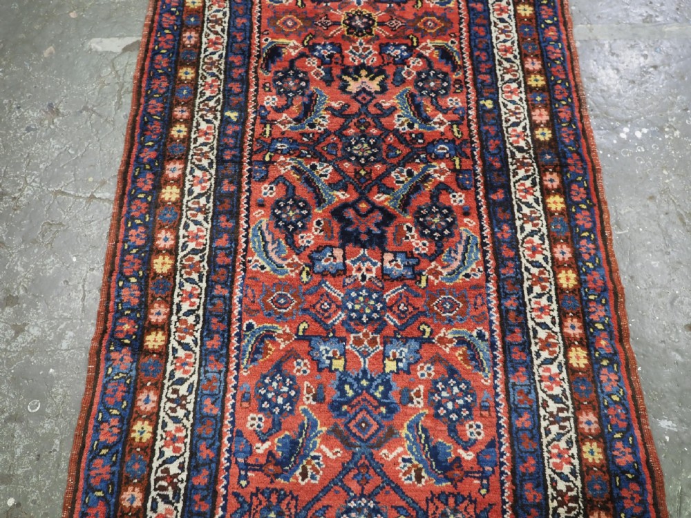 An antique long narrow Persian Kurdish wool runner rug featuring a red central field filled with geometric floral motifs framed by navy blue and cream decorative borders. View VII