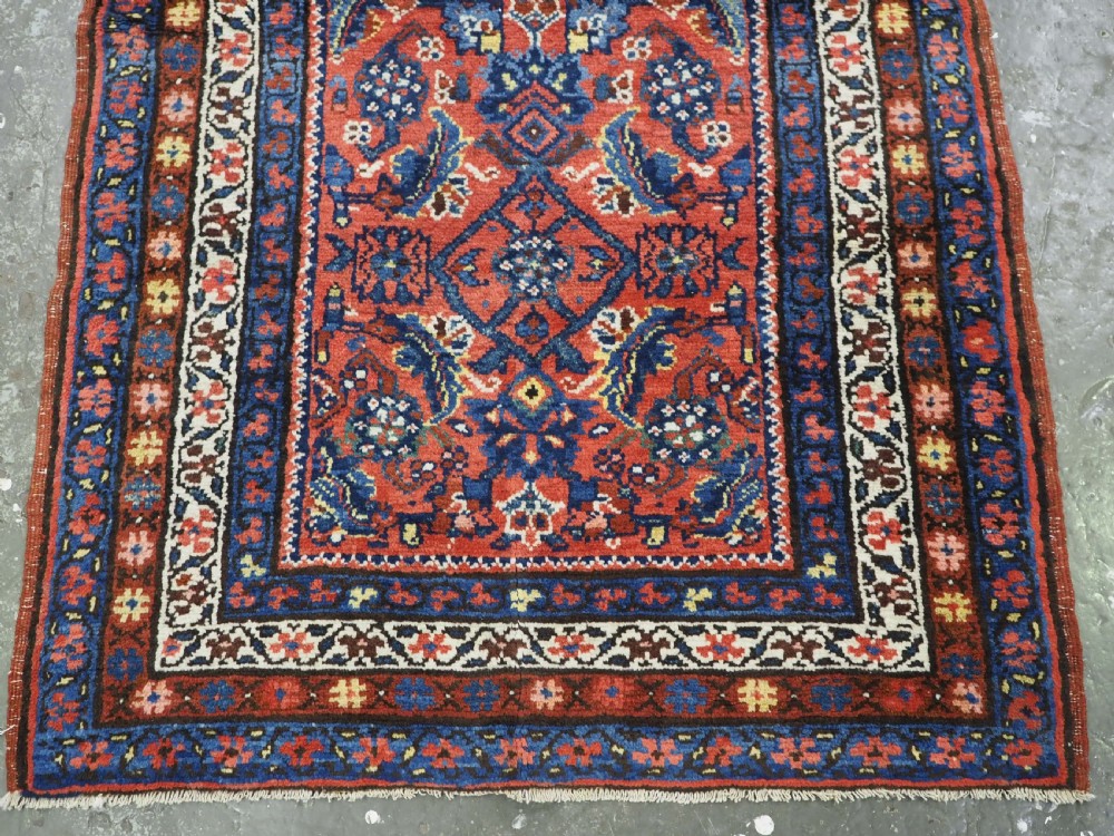 An antique long narrow Persian Kurdish wool runner rug featuring a red central field filled with geometric floral motifs framed by navy blue and cream decorative borders. View VIII