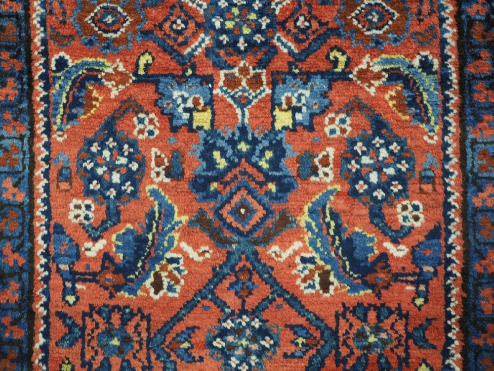 An antique long narrow Persian Kurdish wool runner rug featuring a red central field filled with geometric floral motifs framed by navy blue and cream decorative borders. View IX