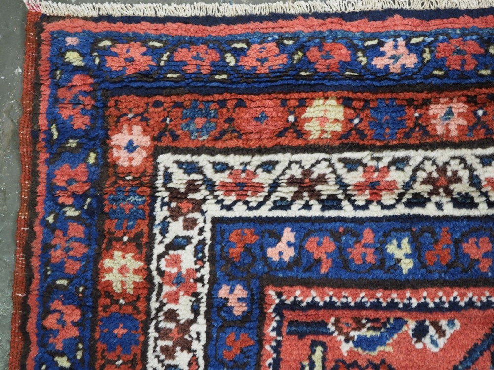 An antique long narrow Persian Kurdish wool runner rug featuring a red central field filled with geometric floral motifs framed by navy blue and cream decorative borders. View X