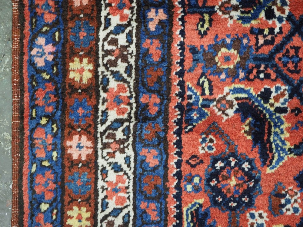 An antique long narrow Persian Kurdish wool runner rug featuring a red central field filled with geometric floral motifs framed by navy blue and cream decorative borders. View XI
