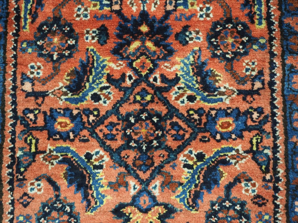 An antique long narrow Persian Kurdish wool runner rug featuring a red central field filled with geometric floral motifs framed by navy blue and cream decorative borders. View XII