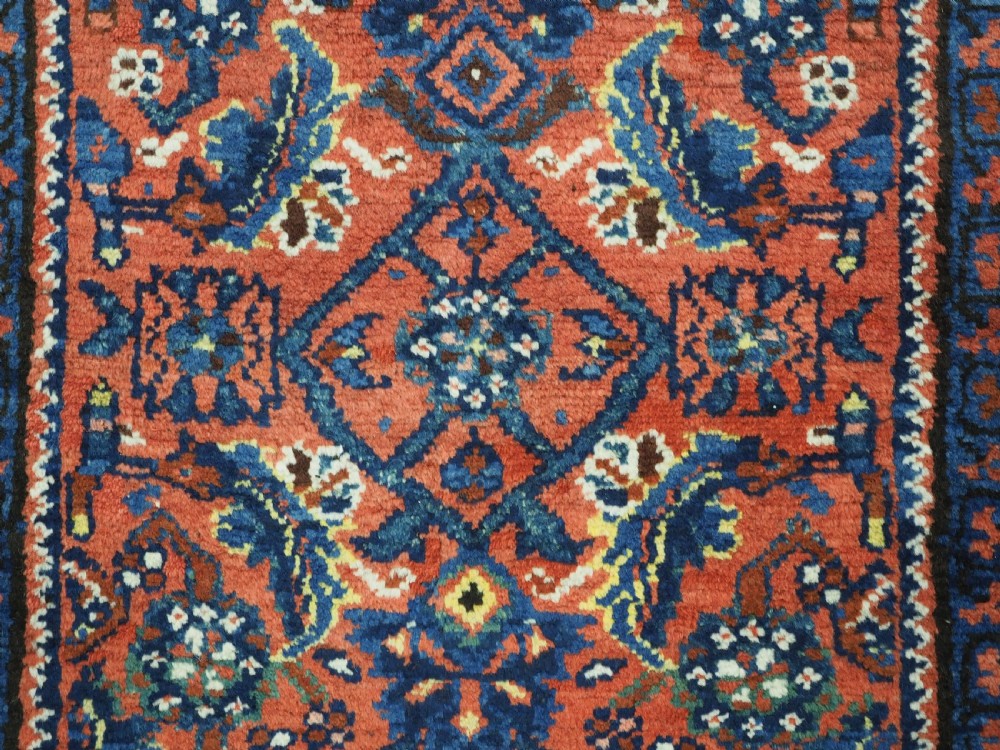 An antique long narrow Persian Kurdish wool runner rug featuring a red central field filled with geometric floral motifs framed by navy blue and cream decorative borders. View XIII
