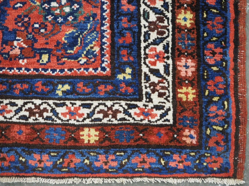 An antique long narrow Persian Kurdish wool runner rug featuring a red central field filled with geometric floral motifs framed by navy blue and cream decorative borders. View XIV