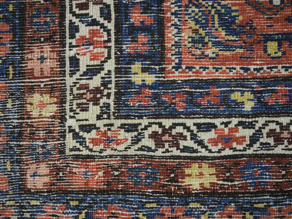 An antique long narrow Persian Kurdish wool runner rug featuring a red central field filled with geometric floral motifs framed by navy blue and cream decorative borders. View XV