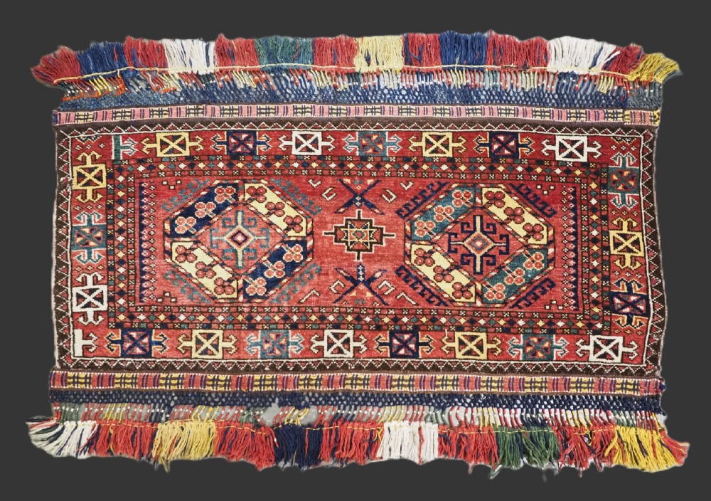 A small rectangular Central Asian wool weaving featuring geometric Turkmen gol motifs in blue and yellow on a red ground with multi-coloured fringe borders.