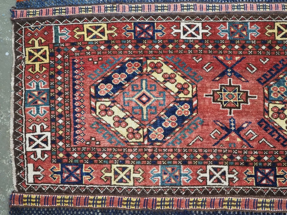 A small rectangular Central Asian wool weaving featuring geometric Turkmen gol motifs in blue and yellow on a red ground with multi-coloured fringe borders. View II