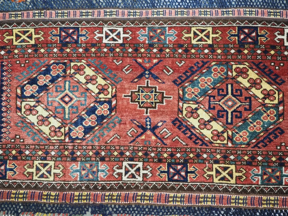 A small rectangular Central Asian wool weaving featuring geometric Turkmen gol motifs in blue and yellow on a red ground with multi-coloured fringe borders. View III