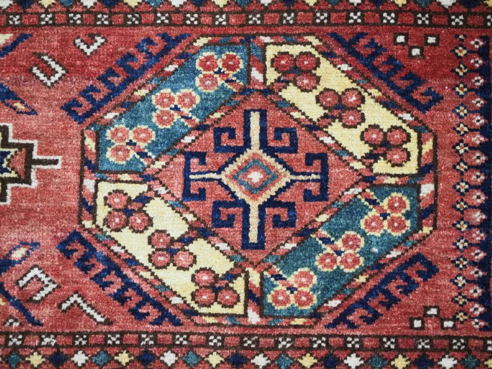 A small rectangular Central Asian wool weaving featuring geometric Turkmen gol motifs in blue and yellow on a red ground with multi-coloured fringe borders. View V