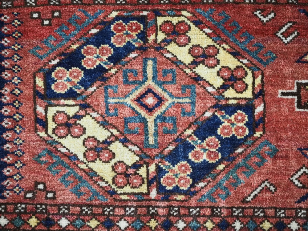 A small rectangular Central Asian wool weaving featuring geometric Turkmen gol motifs in blue and yellow on a red ground with multi-coloured fringe borders. View VI