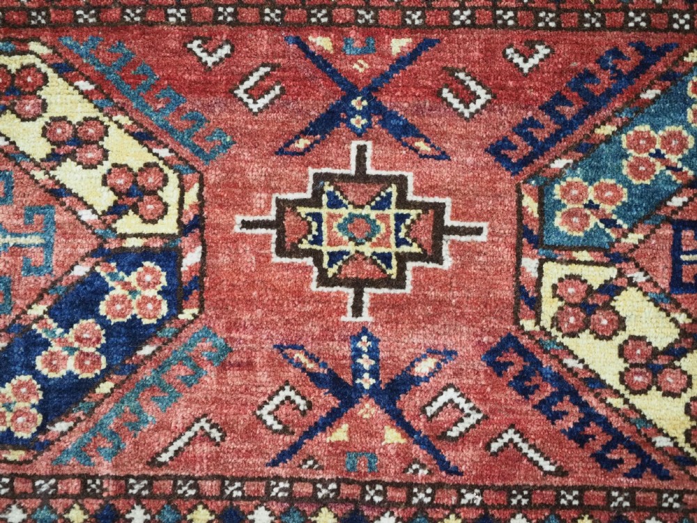 A small rectangular Central Asian wool weaving featuring geometric Turkmen gol motifs in blue and yellow on a red ground with multi-coloured fringe borders. View VII