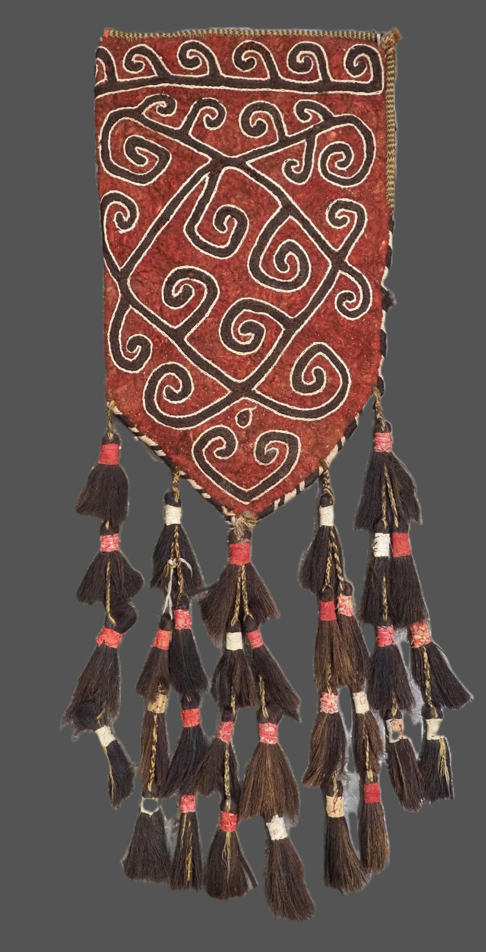 A terracotta red felt panel embroidered with off-white and dark brown scroll motifs, featuring cascading tier tassels made of dark horsehair.