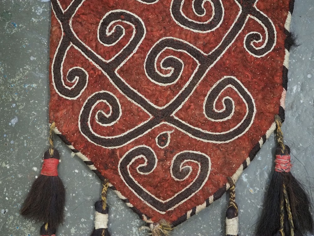 A terracotta red felt panel embroidered with off-white and dark brown scroll motifs, featuring cascading tier tassels made of dark horsehair. View III