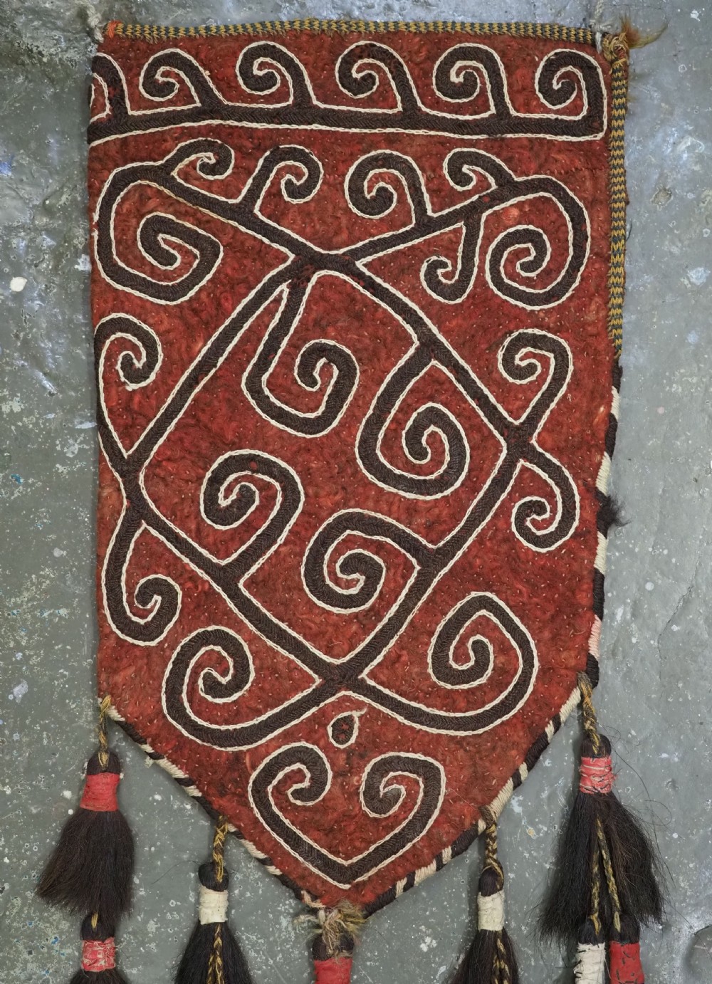 A terracotta red felt panel embroidered with off-white and dark brown scroll motifs, featuring cascading tier tassels made of dark horsehair. View IV