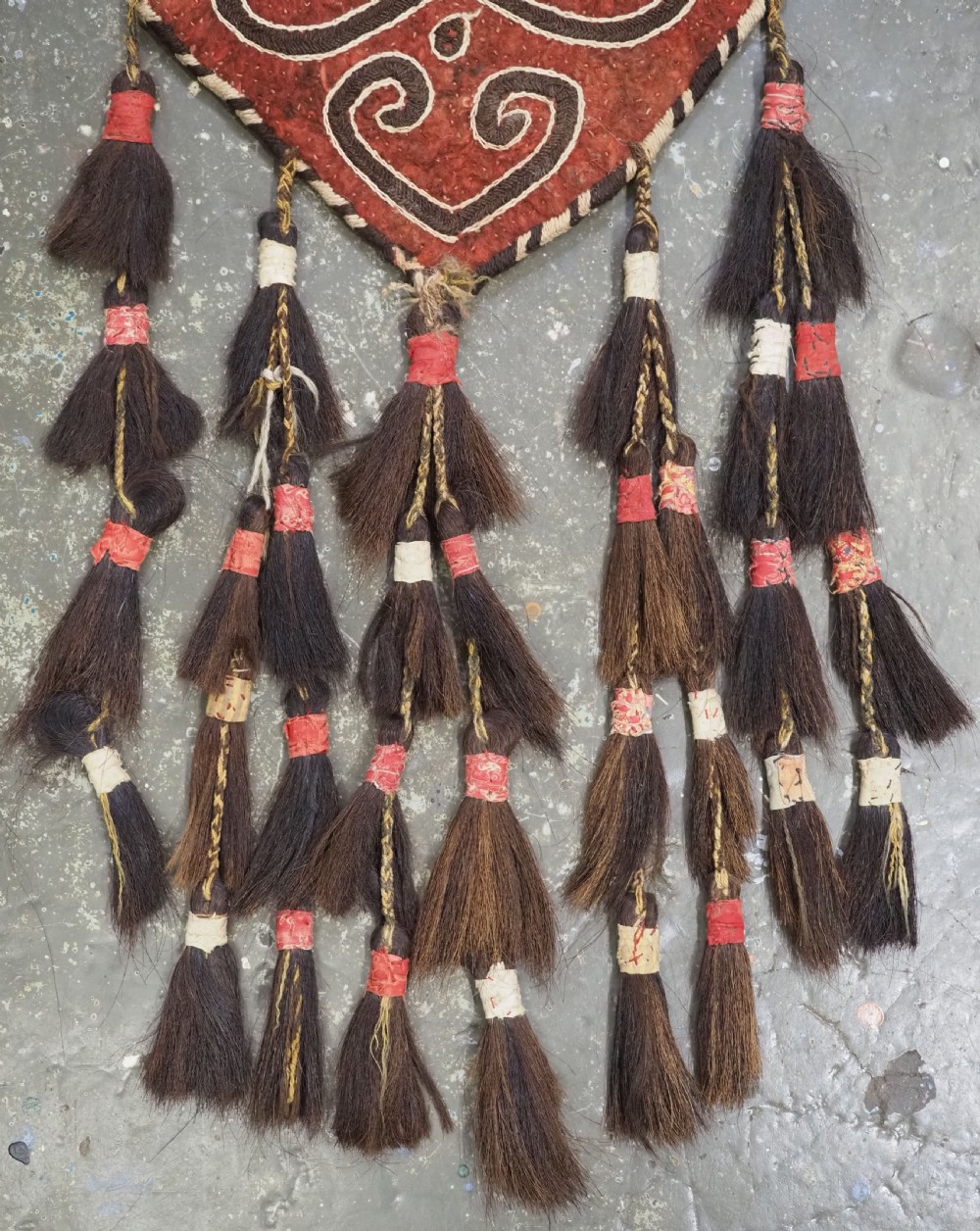 A terracotta red felt panel embroidered with off-white and dark brown scroll motifs, featuring cascading tier tassels made of dark horsehair. View V