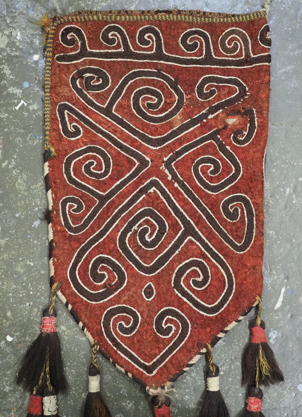A terracotta red felt panel embroidered with off-white and dark brown scroll motifs, featuring cascading tier tassels made of dark horsehair. View VI