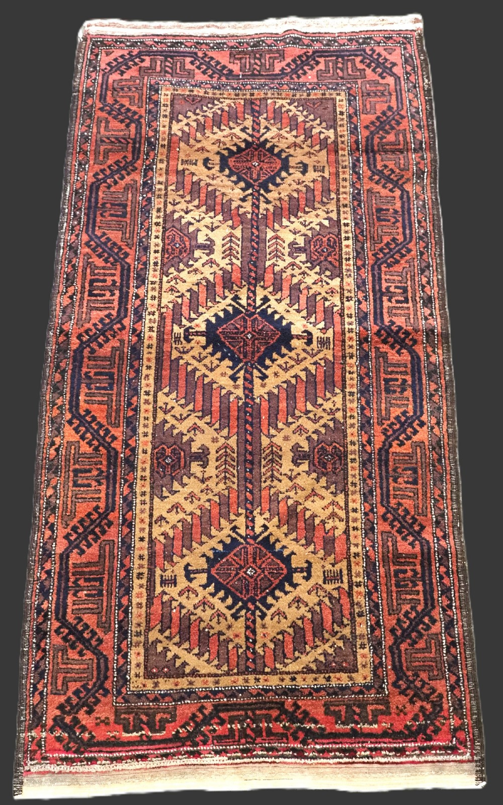 An antique Baluch hand-knotted wool rug featuring three diamond latch-hook medallions on a camel-coloured field with geometric red and navy borders.