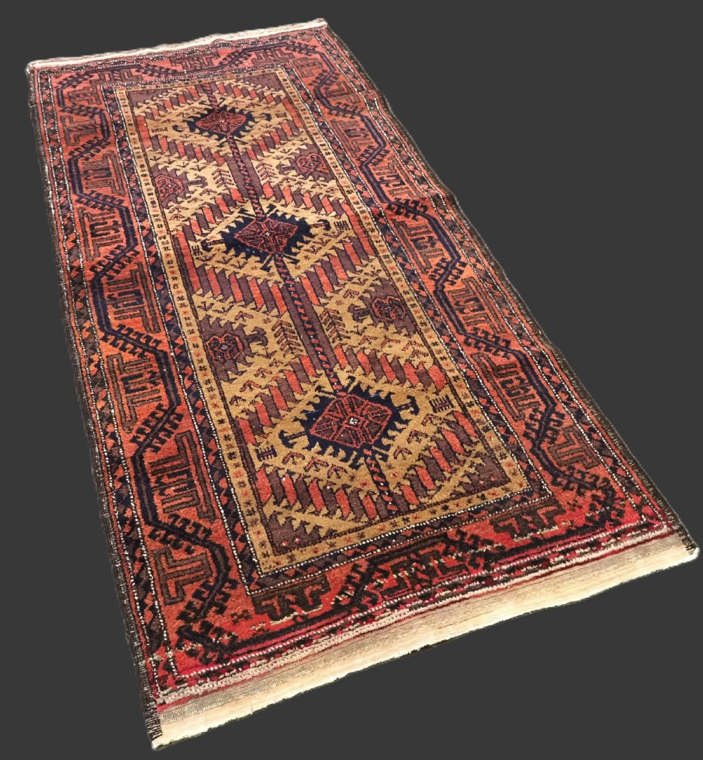 An antique Baluch hand-knotted wool rug featuring three diamond latch-hook medallions on a camel-coloured field with geometric red and navy borders. View II