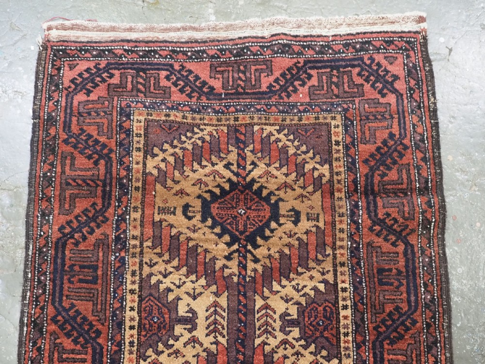 An antique Baluch hand-knotted wool rug featuring three diamond latch-hook medallions on a camel-coloured field with geometric red and navy borders. View III