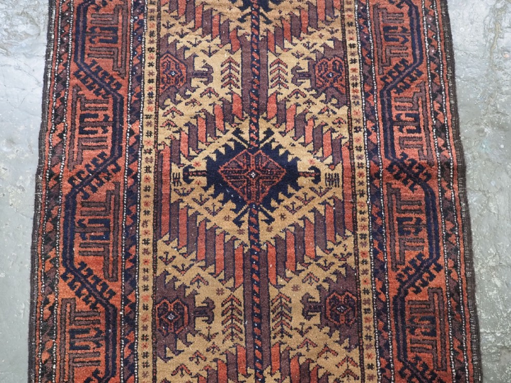 An antique Baluch hand-knotted wool rug featuring three diamond latch-hook medallions on a camel-coloured field with geometric red and navy borders. View IV