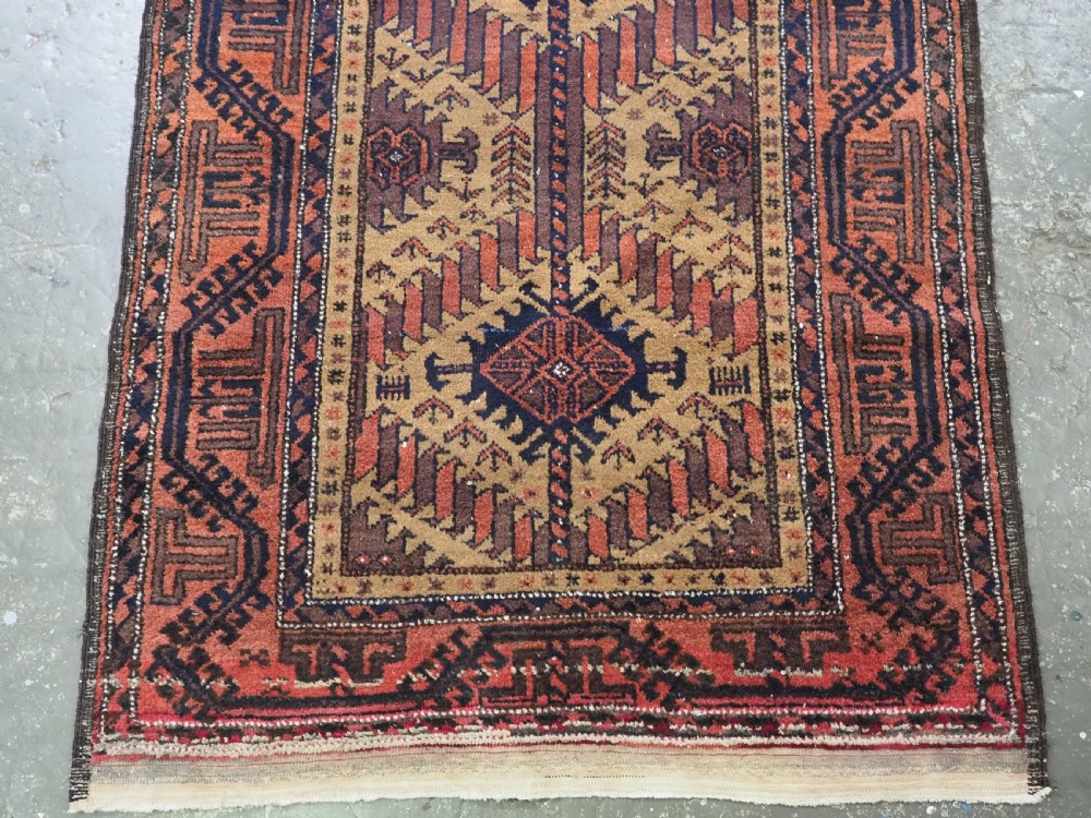 An antique Baluch hand-knotted wool rug featuring three diamond latch-hook medallions on a camel-coloured field with geometric red and navy borders. View V