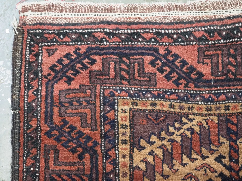 An antique Baluch hand-knotted wool rug featuring three diamond latch-hook medallions on a camel-coloured field with geometric red and navy borders. View VI