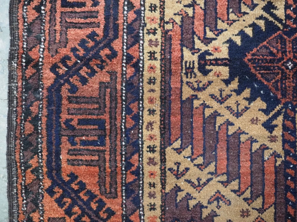 An antique Baluch hand-knotted wool rug featuring three diamond latch-hook medallions on a camel-coloured field with geometric red and navy borders. View VII