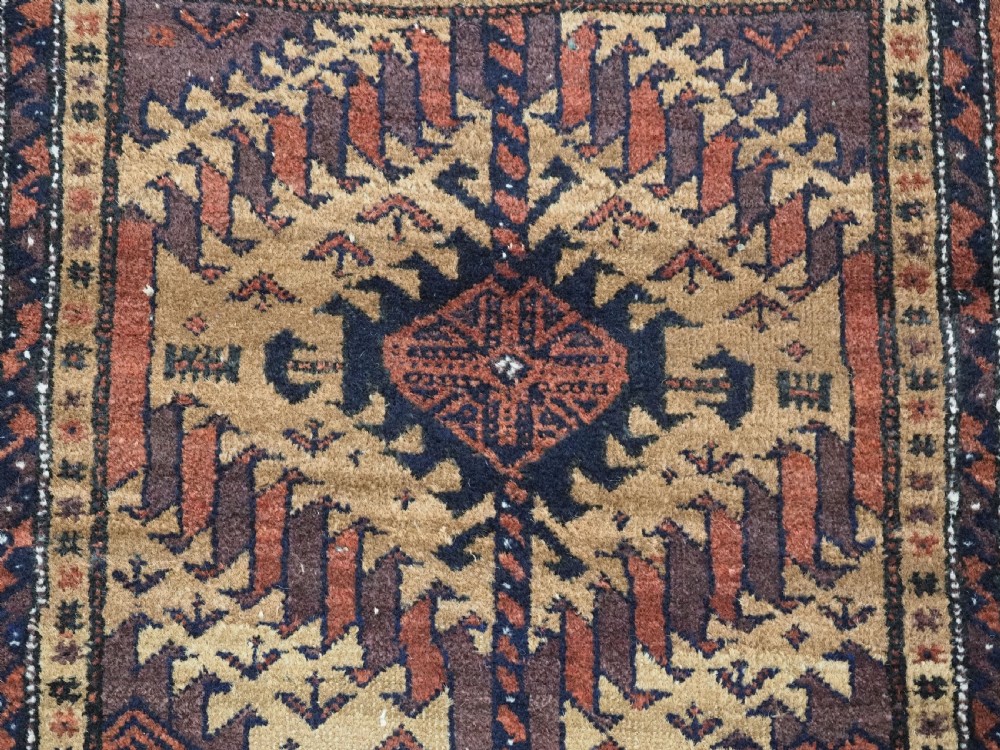 An antique Baluch hand-knotted wool rug featuring three diamond latch-hook medallions on a camel-coloured field with geometric red and navy borders. View VIII