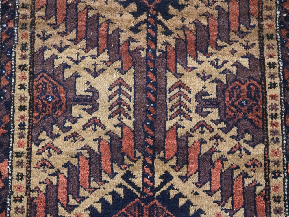 An antique Baluch hand-knotted wool rug featuring three diamond latch-hook medallions on a camel-coloured field with geometric red and navy borders. View IX