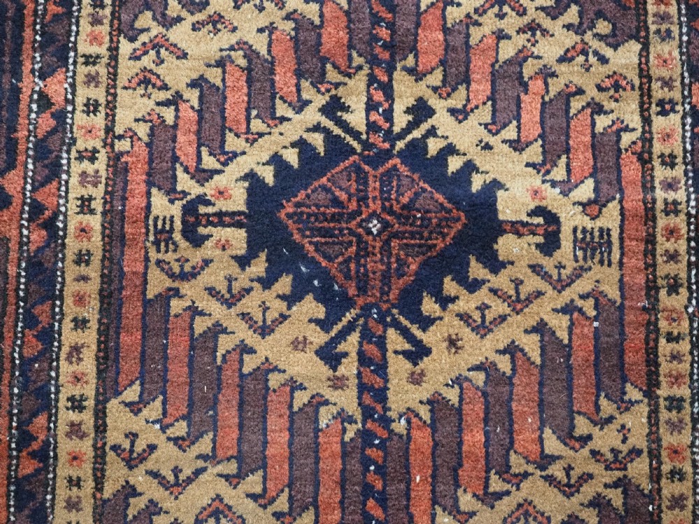 An antique Baluch hand-knotted wool rug featuring three diamond latch-hook medallions on a camel-coloured field with geometric red and navy borders. View X