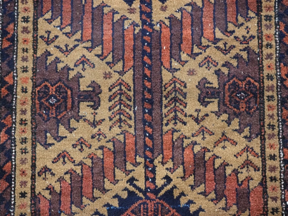 An antique Baluch hand-knotted wool rug featuring three diamond latch-hook medallions on a camel-coloured field with geometric red and navy borders. View XI